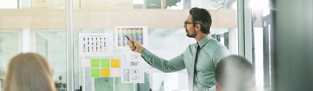 An image of a man in a green button-down shirt standing next to a clear glass window. He is pointing to papers that are posted on the window, which show charts and business plans. 